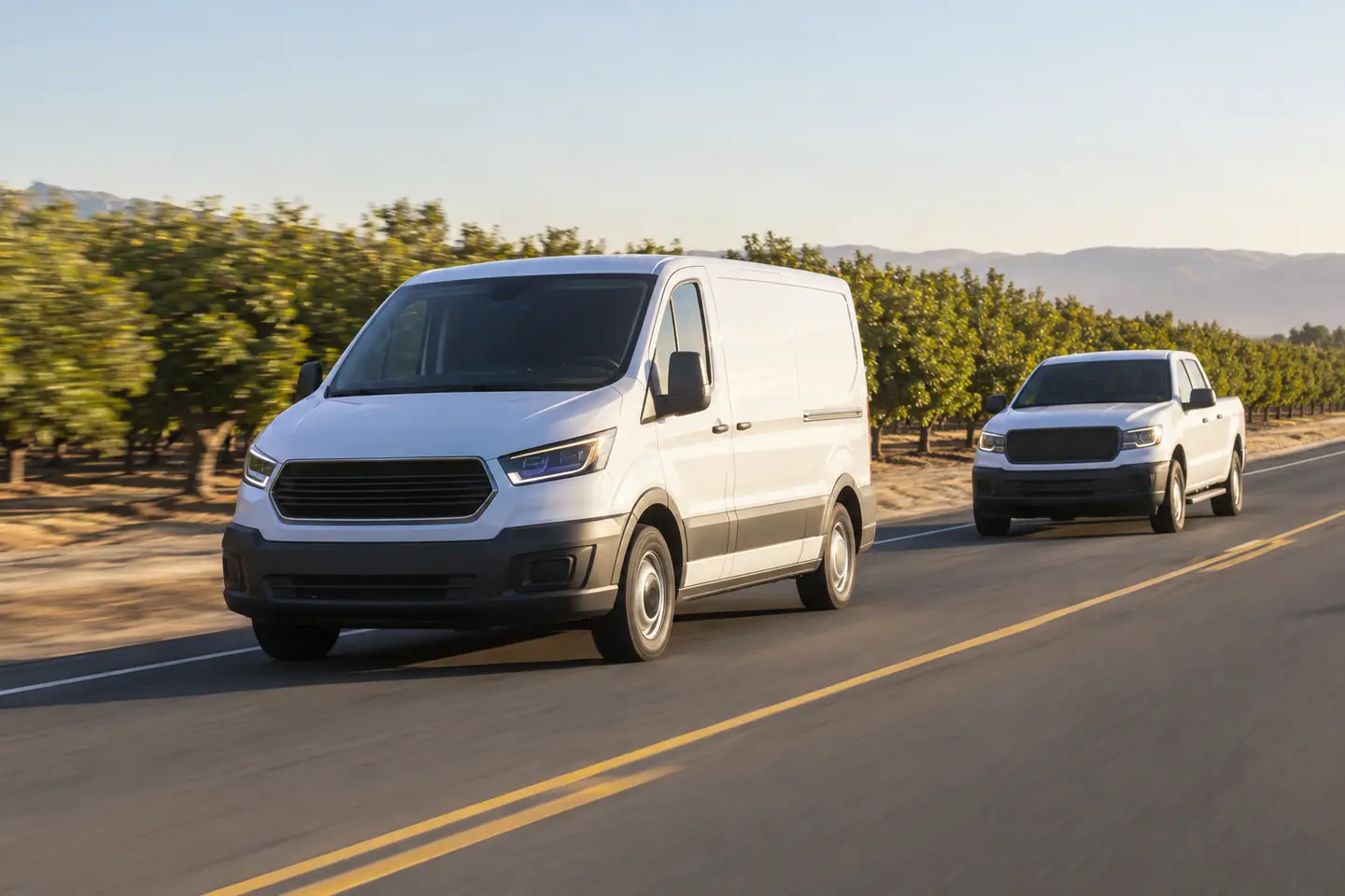 A white cargo van and a pickup truck drive down a road beside an orchard