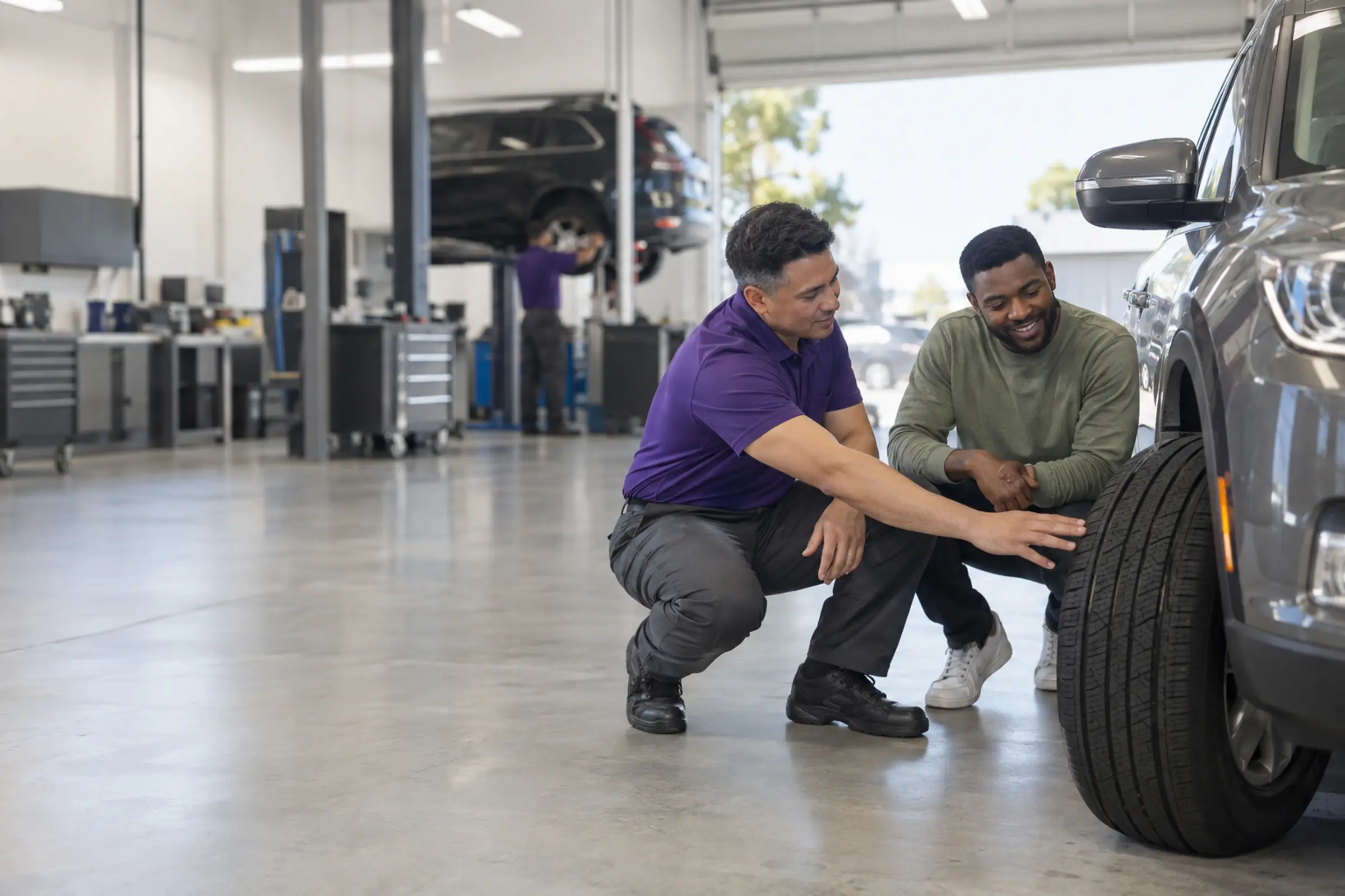 A technician kneels beside a customer’s SUV to show him his tire’s tread